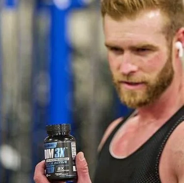 A bearded man in a tank top at a gym holding the DIM 3X bottle, an earbud in one ear; the label is out of focus