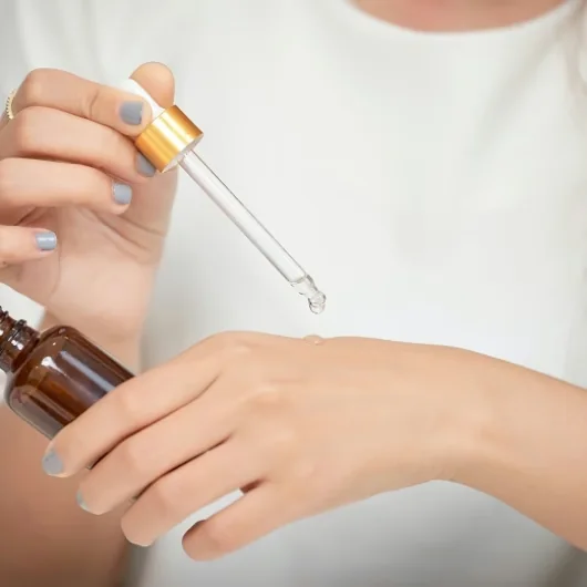 Hand holding a serum dropper above the back of the other hand, dispensing a drop