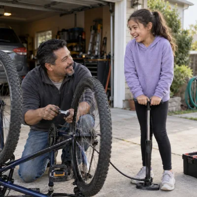 A man in his mid forties kneeling by an upturned bicycle in a driveway, wrench in hand, laughing with his ten-year-old daughter who holds a tyre pump