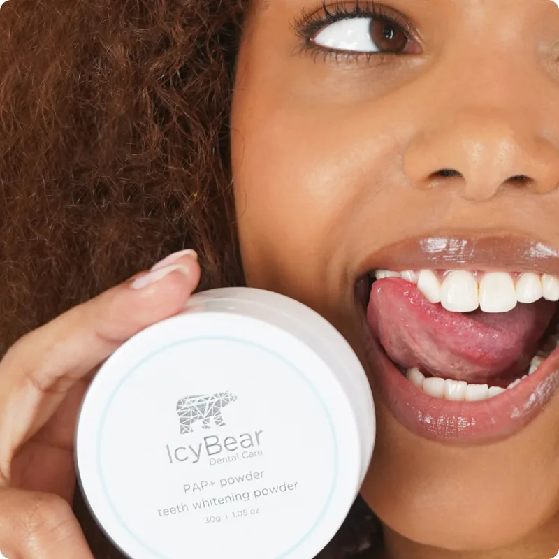 A woman with curly hair smiles and holds the PAP+ Teeth Whitening Powder jar beside her face, manufacturer's photo
