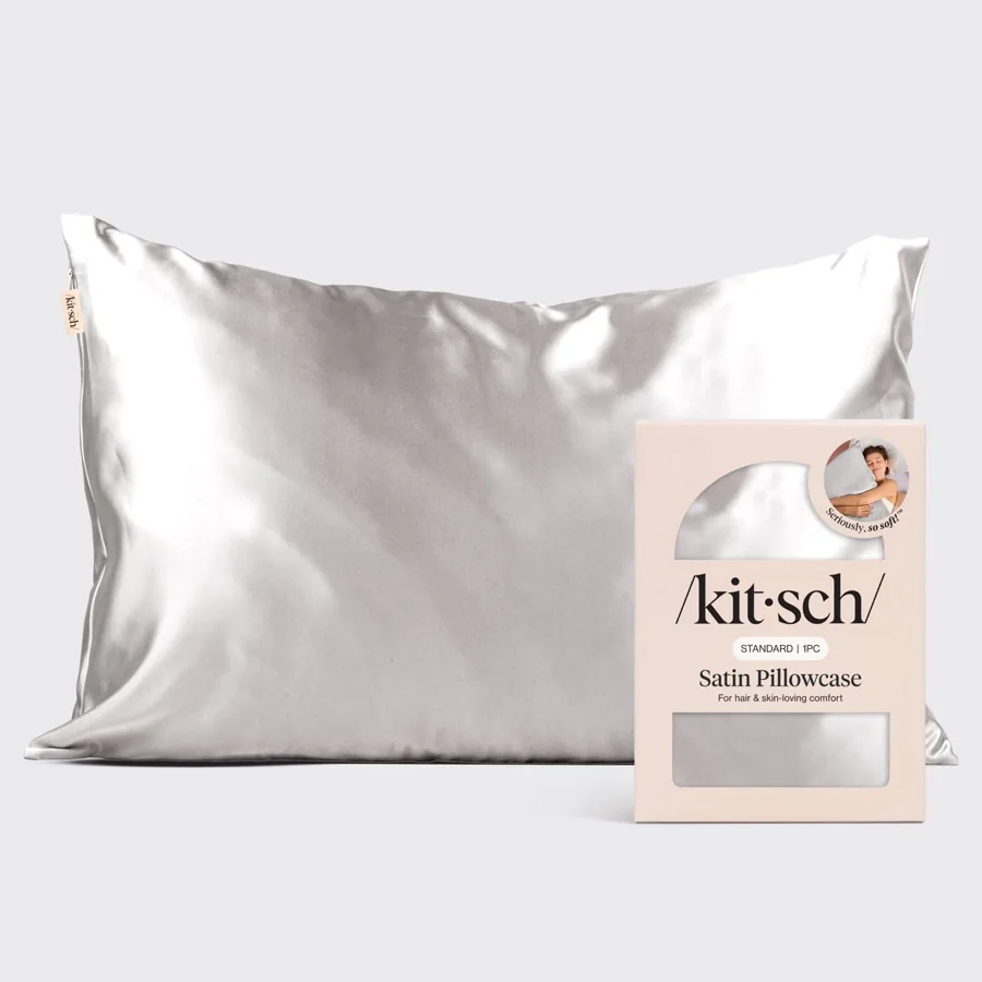 The Kitsch Satin Pillowcase in Silver on a plain background, next to the pale pink retail box it ships in, whose printed lines read For hair and skin-loving comfort and Seriously, so soft