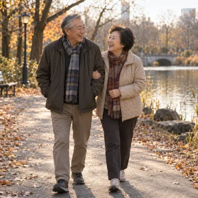 A couple in their late sixties walking arm in arm along a park path in autumn, both laughing