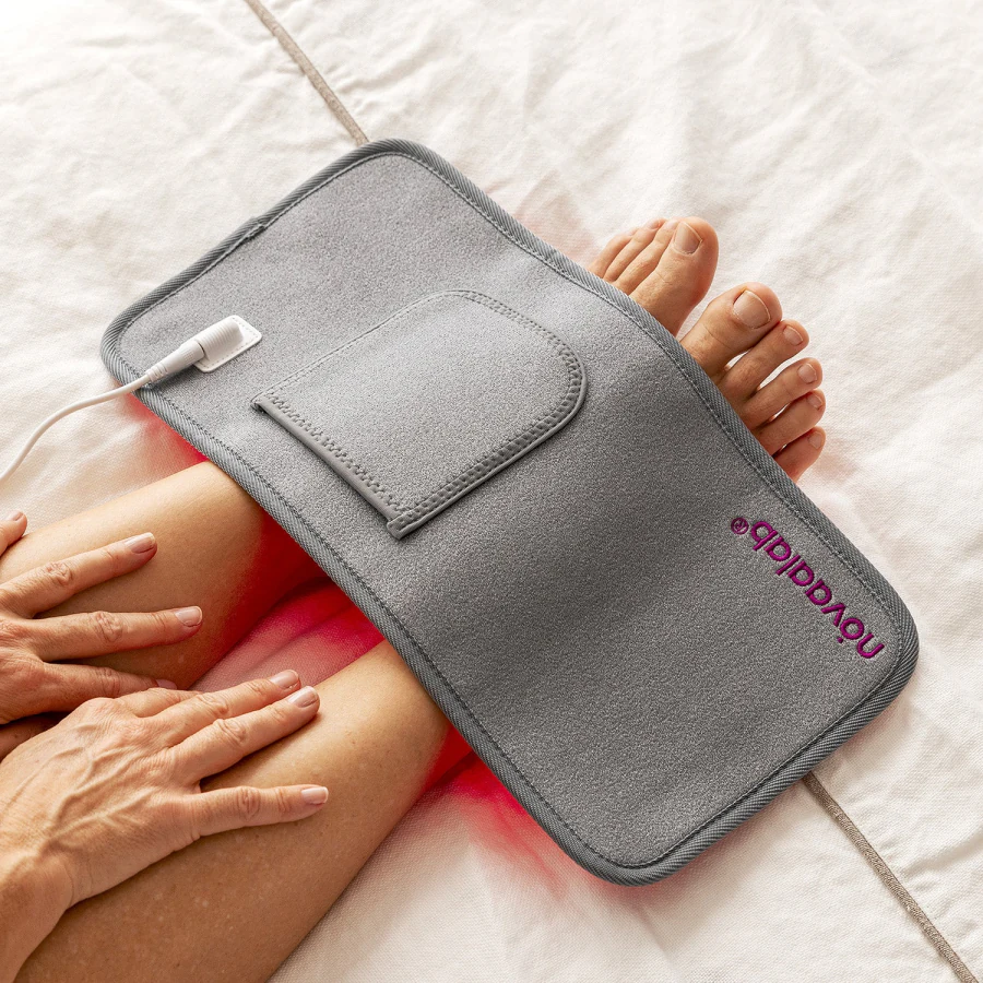 The Light Pad lying face down over a knee on a bed, showing its grey outer shell, the front pocket, the white cable connector and the novaalab logo, with red light escaping at the edges