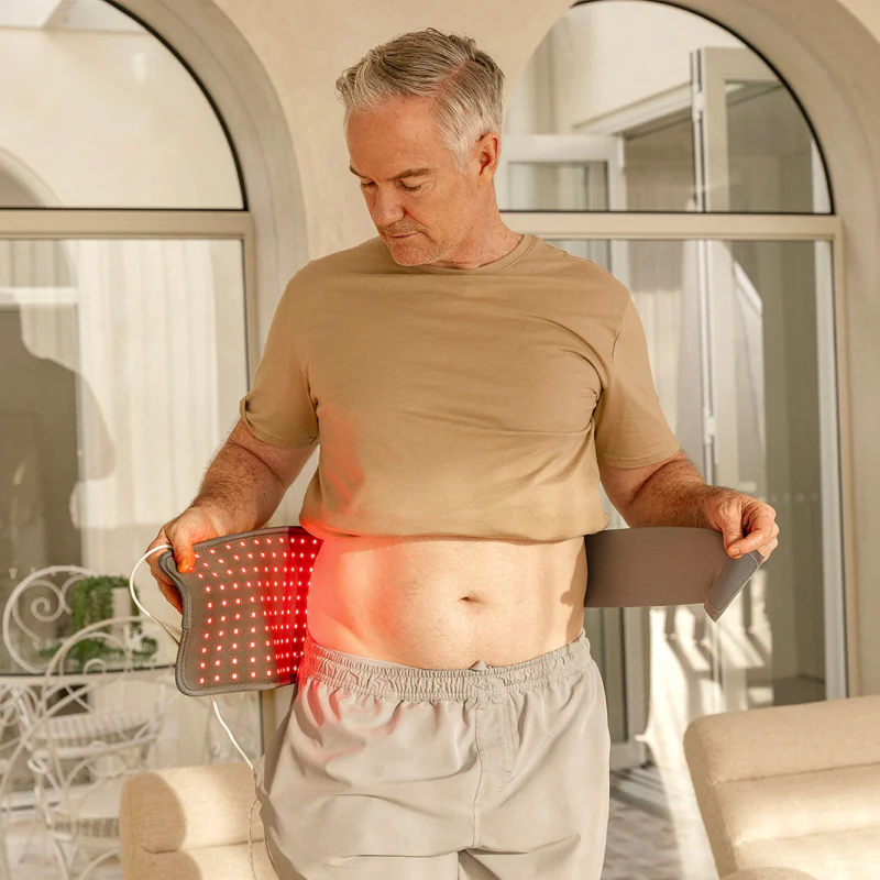 A grey-haired man standing indoors holding the lit Light Pad against his waist, the red light spilling onto his skin