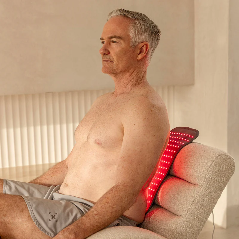 A grey-haired man sitting back in a cream armchair with the lit Light Pad placed between his lower back and the cushion