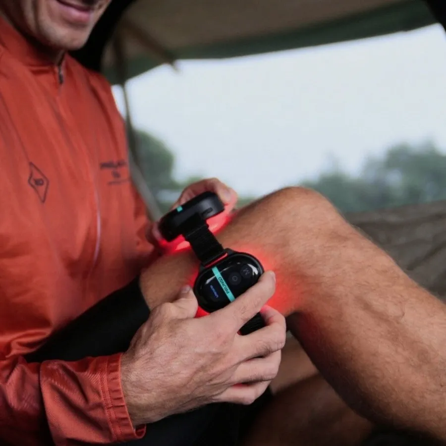 A man in an orange cycling jacket sitting outdoors, strapping a lit PRUNGO module around his knee, red light spilling across the joint