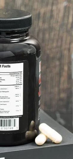 Testosil bottle standing on a grey bench against a wooden background, turned to show the printed label with its ingredient table and barcode, two white capsules beside it